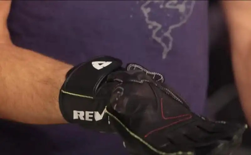 The image provides a close-up view of a man wearing REV’IT! Jerez 3 motorcycle gloves. The gloves are predominantly black with white accents and prominent Rev'it branding. They are made of leather or a similar protective material. The gloves have short cuffs and Velcro straps for adjustability. The focus of the image is on the gloved hands gripping the handlebars, showcasing the gloves ventilation.